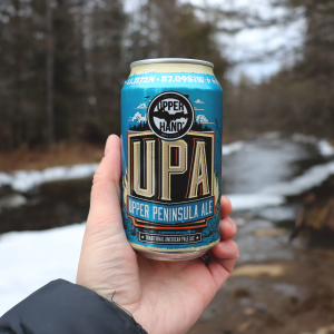 A hand holding a blue can of beer labeled UPA in front of a snowy river"